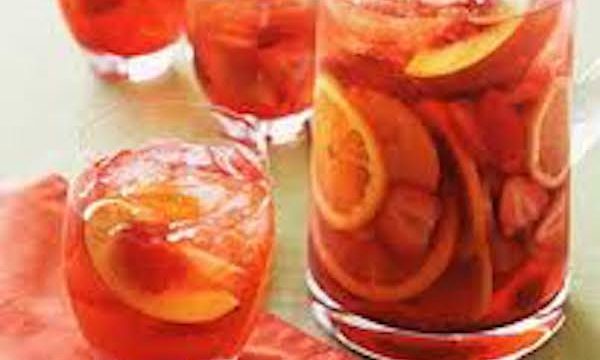 A large glass pitcher and three glasses filled with rose colored sangria and fruit. One of the glasses in the foreground is sitting on a coral napkin.