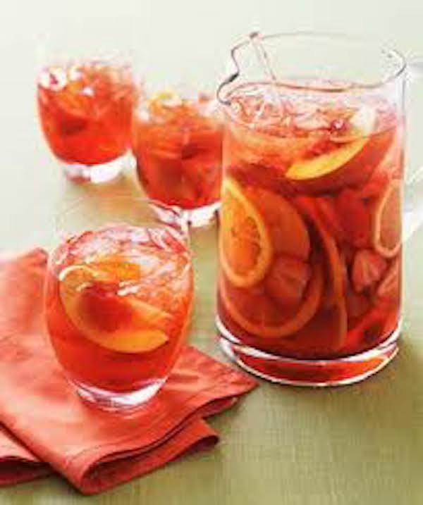 A large glass pitcher and three glasses filled with rose colored sangria and fruit. One of the glasses in the foreground is sitting on a coral napkin.