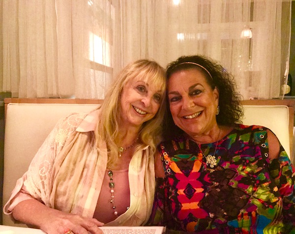 A blond woman and a dark haired woman with heads touching are seated in a booth with a curtain behind them.