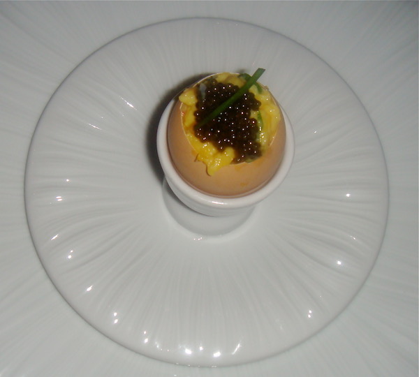 Small white plate on large white plate in center is an egg cup holding a brown egg shell filled with egg, lobster and topped with caviar