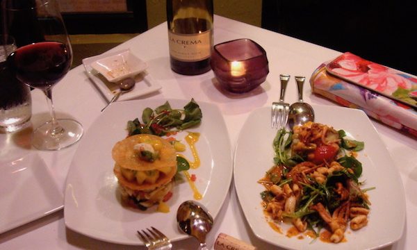 Square table in restaurant with white cloth and two entrees along with bottle of wine and votive