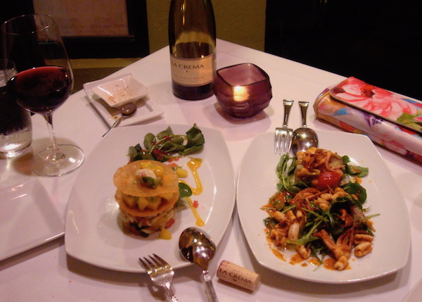 Square table in restaurant with white cloth and two entrees along with bottle of wine and votive