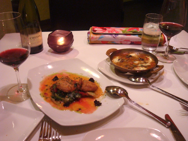 Square table in restaurant with white tablecloth, two plates of appetizers and bottle of wine
