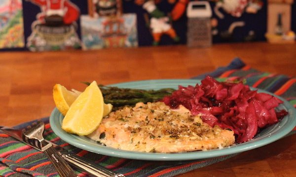 An aqua dinner plate topped with salmon, asparagus, braised red cabbage and two lemon wedges sitting on a butcher block.