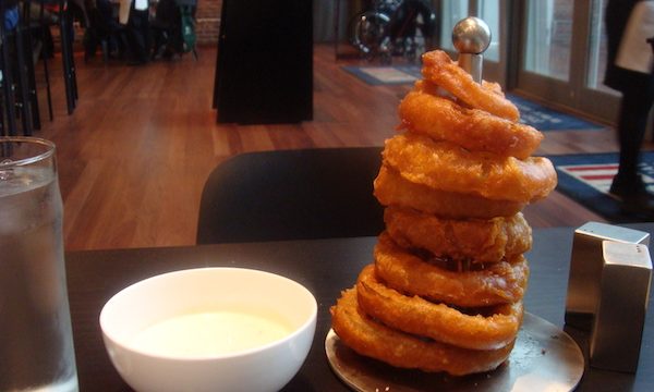 A stack of crispy golden brown onion rings on a dowel with a white bowl of sauce on the left side