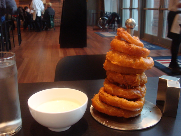 A stack of crispy golden brown onion rings on a dowel with a white bowl of sauce on the left side