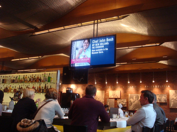 Inside of a restaurant with a TV hanging from the ceiling showing Chef John Besh's picture