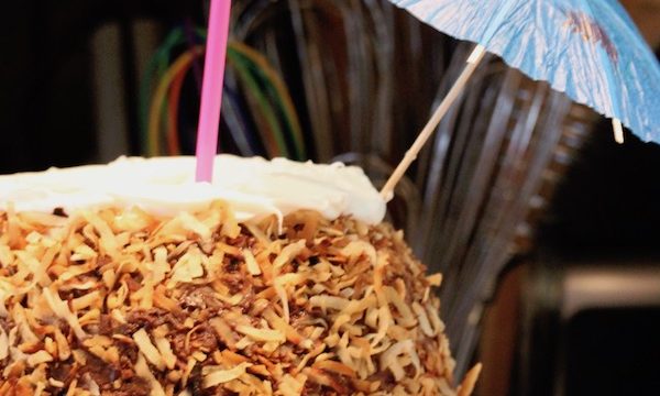 A cake shaped like a pineapple with toasted coconut on the outside, a pink straw and cocktail umbrella on top