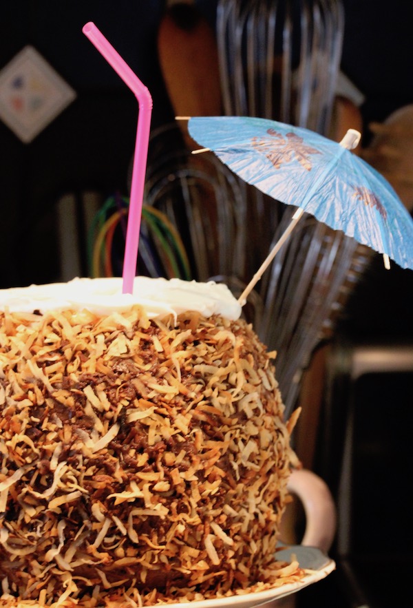 A cake shaped like a pineapple with toasted coconut on the outside, a pink straw and cocktail umbrella on top