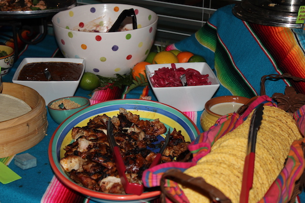 A platter of chicken, tortillas, bowls and condiments sitting on a Mexican blanket