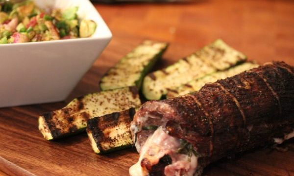 A cooked flank steak roulade, grilled zucchini and a partial white ceramic square bowl with salsa all on a butcher block.