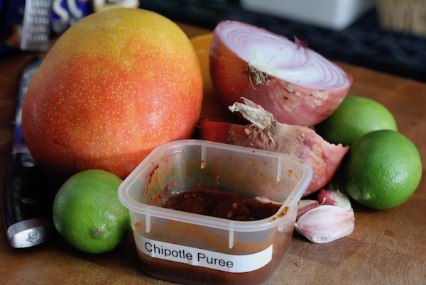 A whole ripe mango, a lime, small square container of Chipotle Purée, a clove of unpeeled garlic and two halves of a red onion sitting on a butcher block.