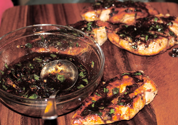 Grilled chicken breasts topped with a dark vinaigrette flecked with scallion slices sitting on a wood cutting board along side a glass bowl with the vinaigrette and a small ladle.
