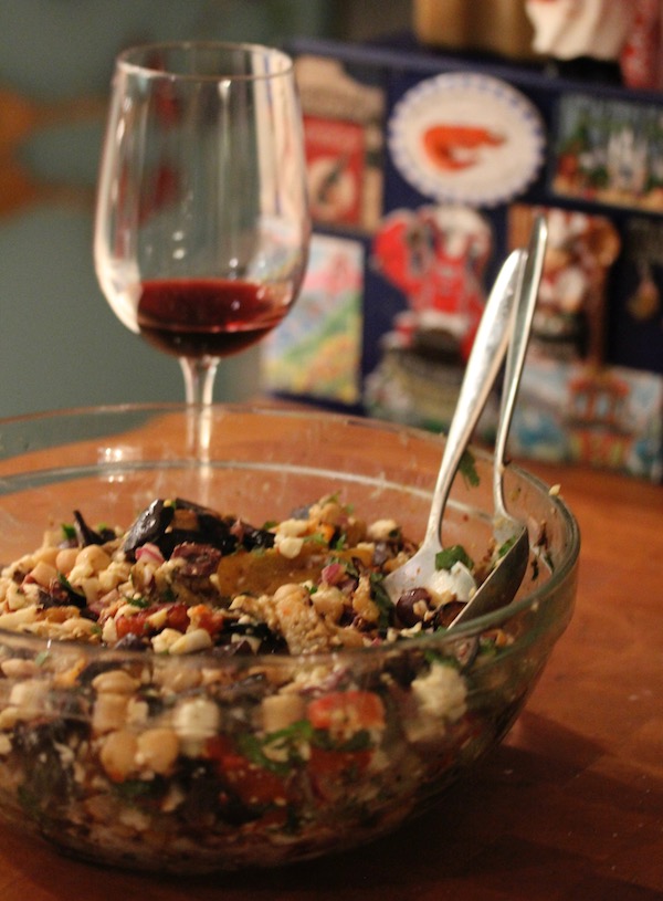 Glass bowl filled with chickpeas, feta, red peppers, red onion, eggplant and black olives with serving utensils sticking up. Bowl is on a butcher block and behind bowl a glass of red wine.
