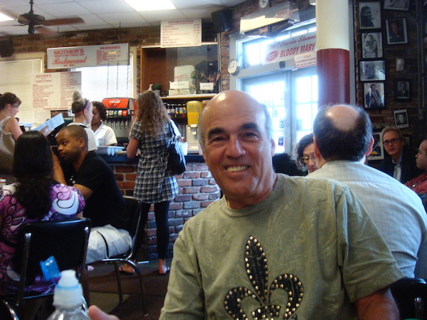 Smiling bald man wearing an olive green t-shirt with a brass studded fleur de lis in a crowded restaurant