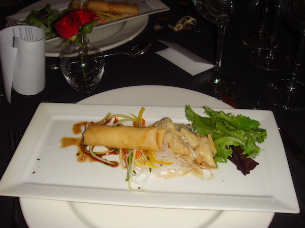 Rectangular white plate atop a white charger. The rectangular plate holds a golden spring roll, julienne vegetables, dumplings and greens
