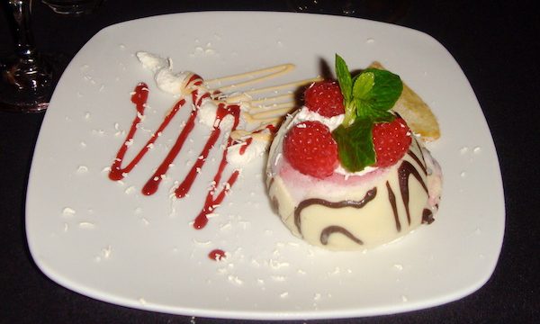 White square plate on black tablecloth on right side of plate is a white chocolate orb topped with 3 raspberries and mint leaves and a squiggle of red raspberry coulis to the left of the orb