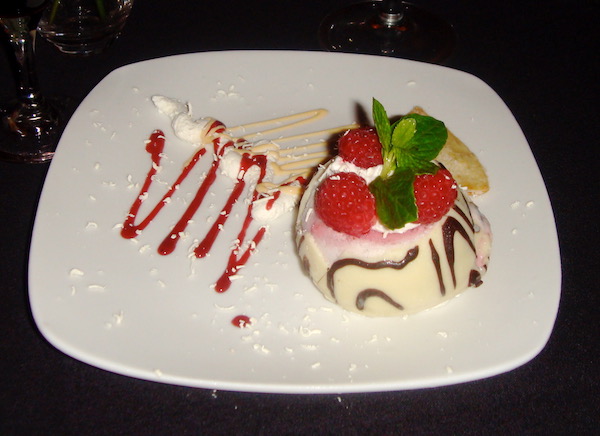 White square plate on black tablecloth on right side of plate is a white chocolate orb topped with 3 raspberries and mint leaves and a squiggle of red raspberry coulis to the left of the orb