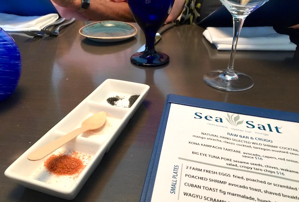 A white divided narrow rectangular plate with three different sea salts and an ivory spoon on a set table next to a menu