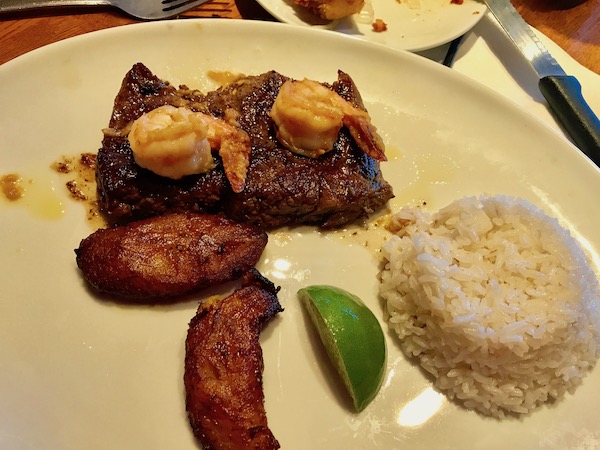 Grilled meat, fried plantains, a scoop of white rice and a lime wedge on a white plate