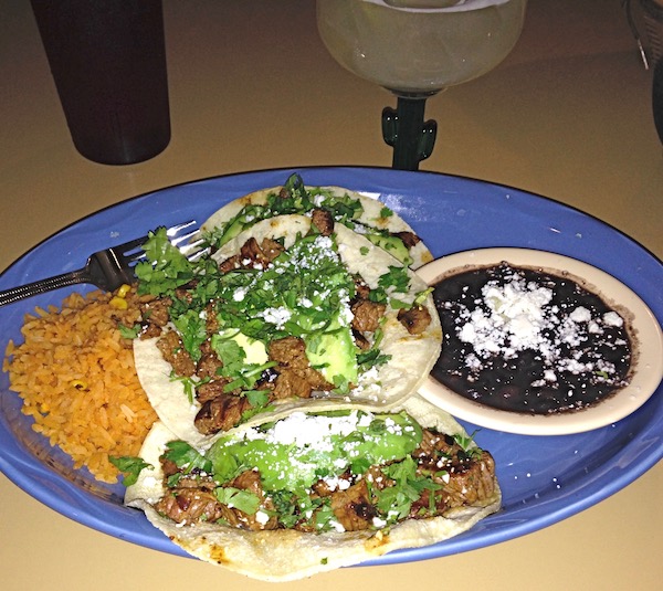 A blue plate topped with tortillas, steak and shredded greens with a dish of black beans and rice.