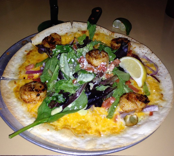 A white plate that is completely covered by a quesadilla and topped with arugula and scallops.