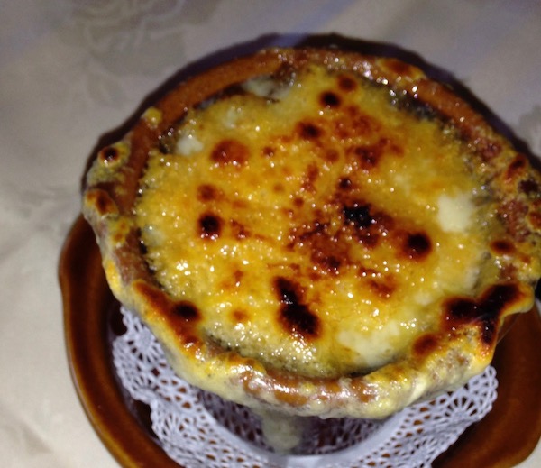 A brown ceramic crock sit-in on a doily on a brown plate. The crock is covered with melted cheese that is brown in spots.