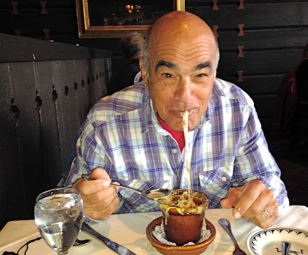 A man in a plaid shirt eating French Onion Soup with melted cheese trailing from crock to his mouth.