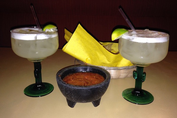 Two filled margarita glasses with a molcajete filled with salsa between them and a basket of chips in the background..
