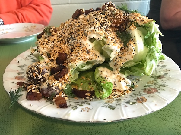 A white plate with a huge wedge of iceberg lettuce covered with bacon, dressing and everything bagel spice.