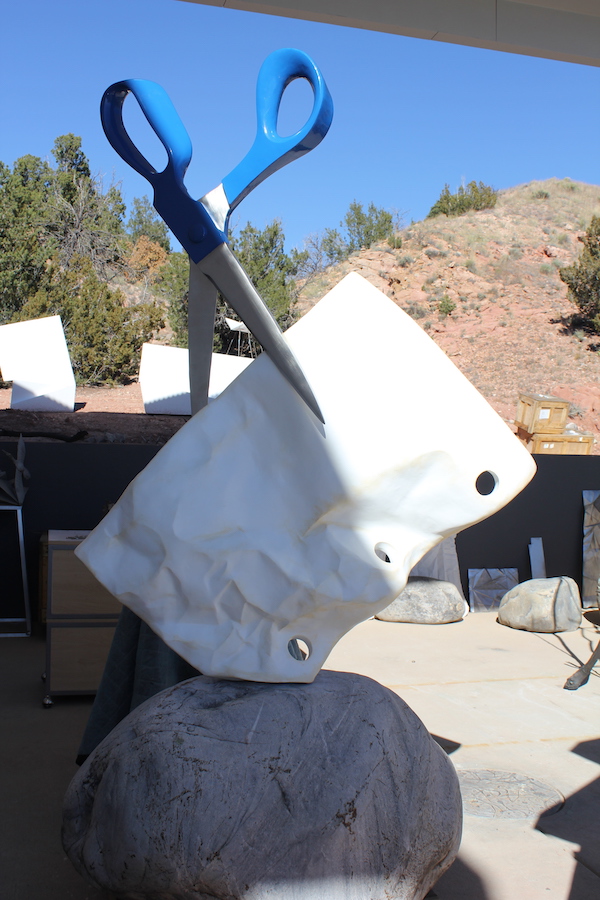A large sculpture of a rock topped with a piece of paper and a large scissors appearing to cut the paper.