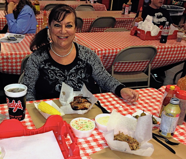 A woman sitting at a picnic bench cover in a red and white checkered cloth. In front of her is a full bbq meal.