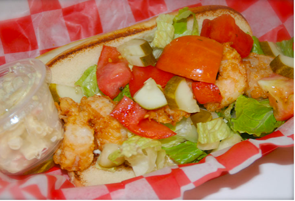 A hotdog bun filled with fried shrimp and tomatoes sitting on red and white checked deli paper
