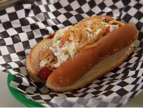 Hot Dog With Coleslaw and Onion Straws in a bun sitting on black and white checked deli paper