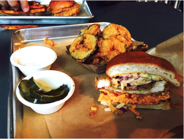 A table with braised greens in a little white dish, fried zucchini slices and a fried chicken sandwich on butcher paper on a sheet pan