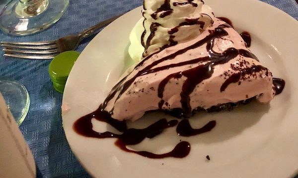 A white plate on a dining table with a slice of pink pie drizzled with chocolate sauce and a large swirl of whipped cream