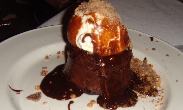 White plate with small chocolate lava cake that has a scoop of vanilla ice cream on top that is drizzled with bittersweet chocolate sauce