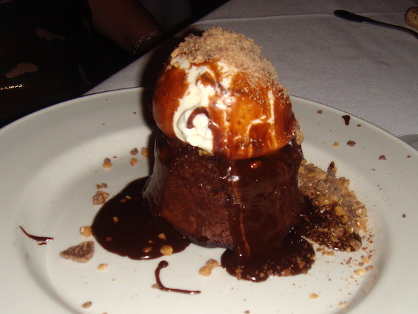 White plate with small chocolate lava cake that has a scoop of vanilla ice cream on top that is drizzled with bittersweet chocolate sauce