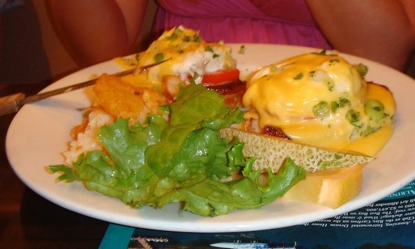 A white plate topped with eggs benedict, home fries, greens and cantaloupe slices