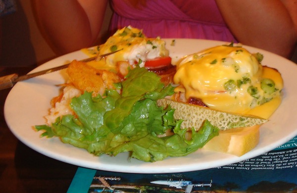 A white plate topped with eggs benedict, home fries, greens and cantaloupe slices