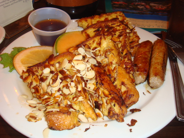 A white plate topped with triangles of almond covered French toast, two sausage links, fruit slices and a small plastic container of maple syrup