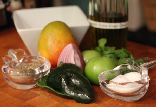 A butcher block with salt and pepper dishes, poblano pepper, mango and tomatillos with a square white bowl behind the mango