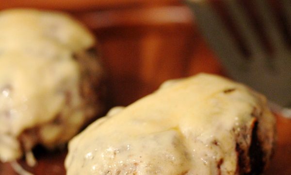 Two burgers with melted cheese covering the top and dripping down the sides, resting on a cutting board