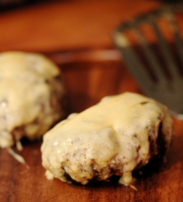 Two burgers with melted cheese covering the top and dripping down the sides, resting on a cutting board
