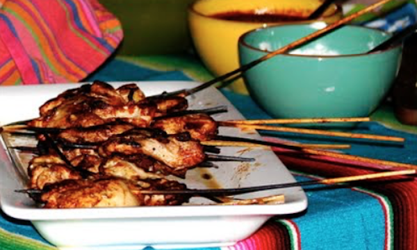 A white platter filled with chicken on bamboo skewers with a yellow bowl and turquoise bowl in the upper right