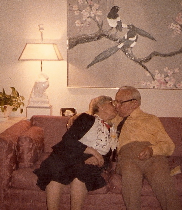 An older man and woman sitting on a mauve sofa kissing