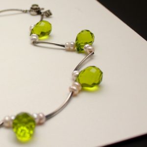 Partial view of a necklace of teardrop shaped faceted peridots, freshwater pears and sterling silver curved tubes on a white board.