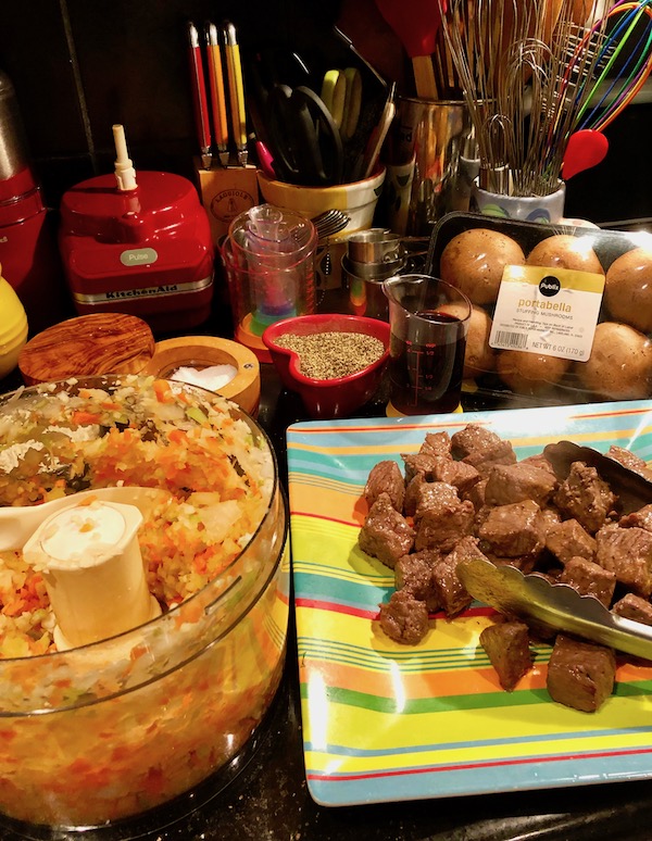 On the left a food processor hold chopped carrots, celery, onion and garlic. On the right a colorful striped plate is topped with cubes of seared beef.