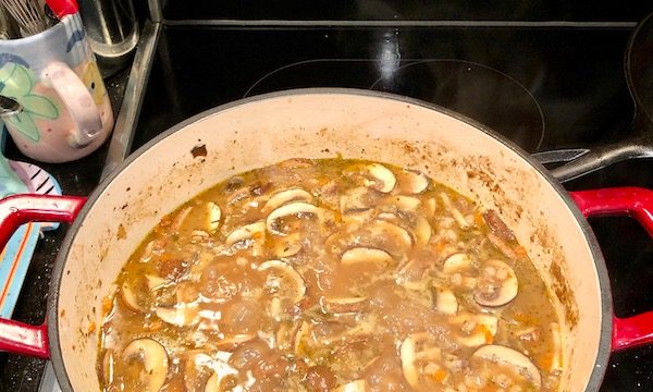 An enameled cast iron Dutch oven with red handles filled with all of the ingredients of Beef, Barley and Portobello Mushroom soup that is cooked to perfection.