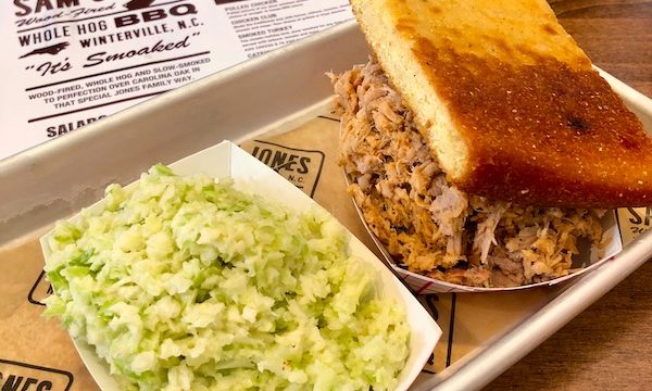 Sitting atop a menu from Sam Jones BBQ is a quarter sheet tray holding chopped pork, a slice of toasted cornbread and a paper container of coleslaw.